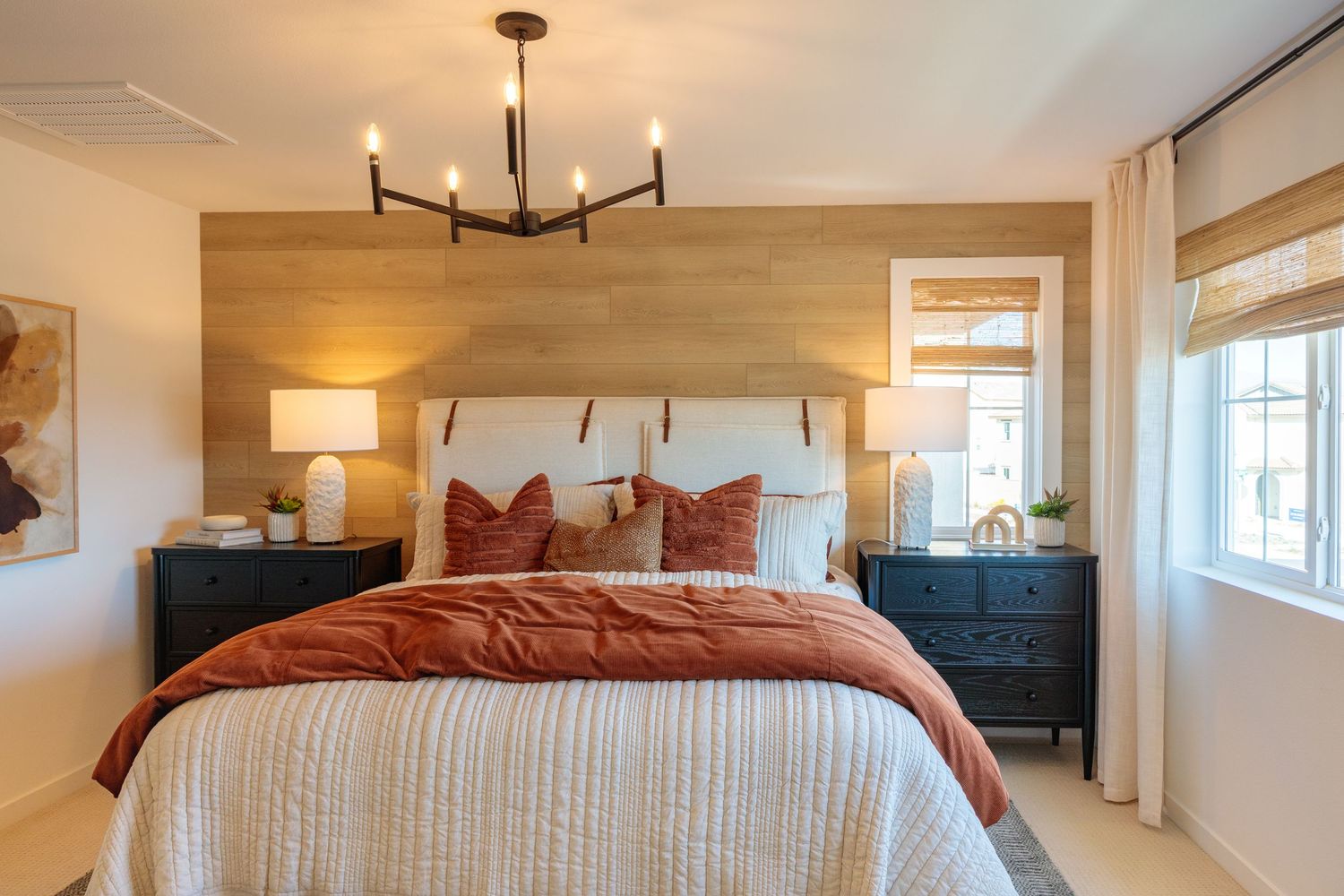 A modern bedroom features a rustic chandelier, white walls, woven window shades and earth-toned bedding.