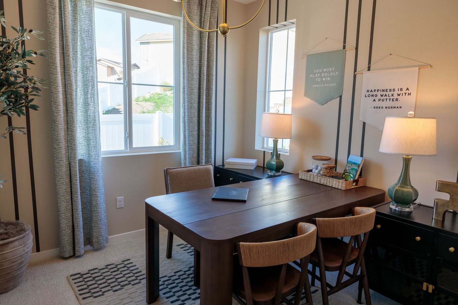 Modern home office with dark wood desk, upholstered chairs, tall windows with floor-length curtains, and decorative lamps.