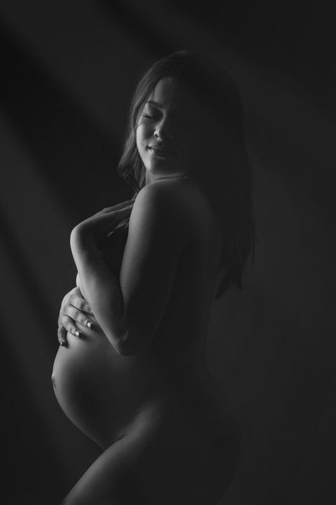 Black and white maternity portrait of a pregnant woman cradling her baby bump in soft dramatic lighting.