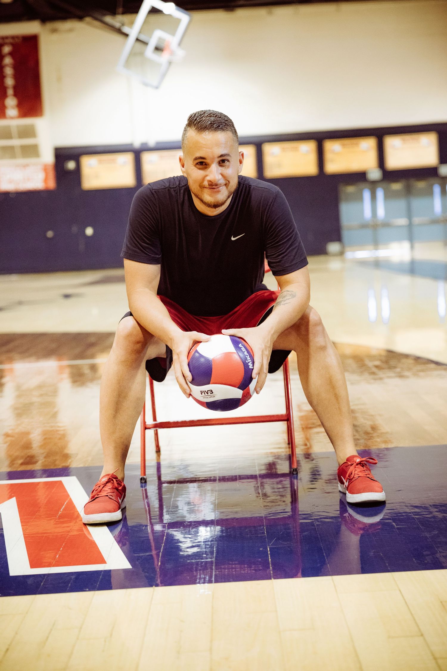 Man in black Nike shirt sits on a chair holding a volleyball on an indoor gym court.