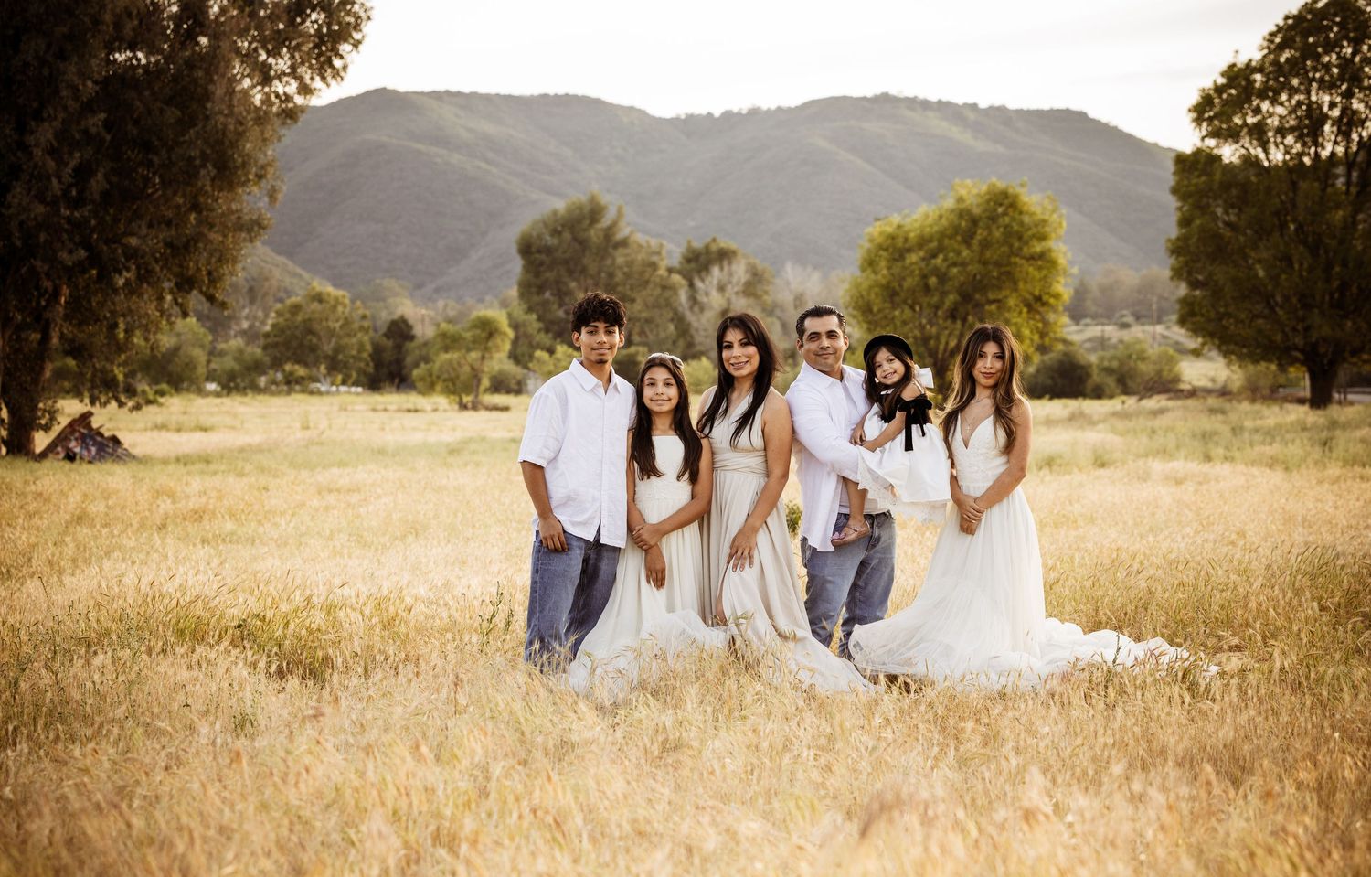 Family of six posing in golden field with mountain backdrop, dressed in white outfits for a professional outdoor photo session.