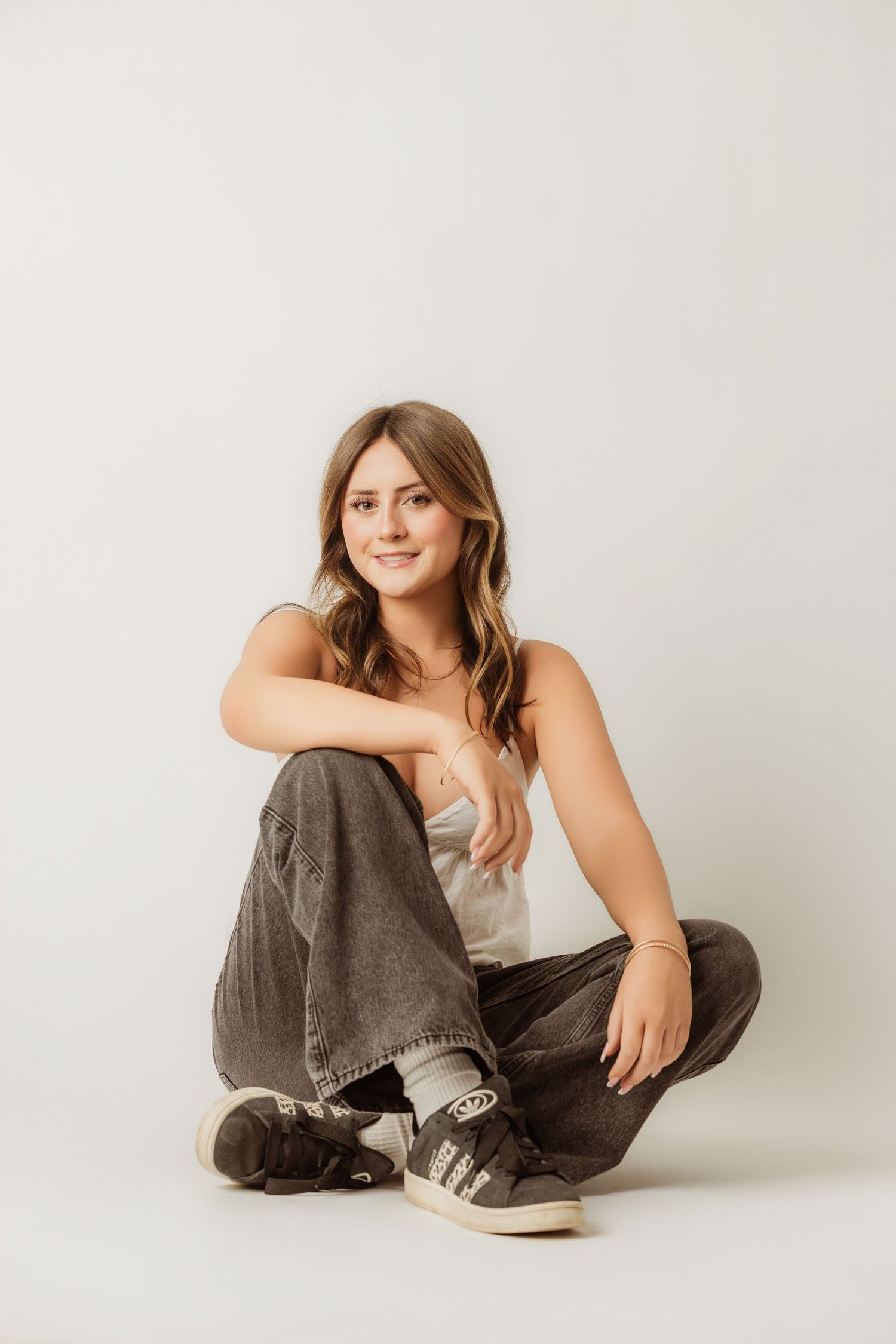 Young woman sitting cross-legged on white background in casual outfit.