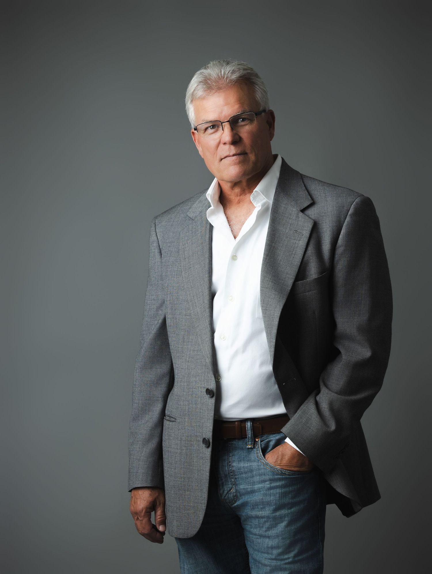 Professional portrait of a person in a gray blazer, white shirt and jeans against a dark gray studio backdrop.