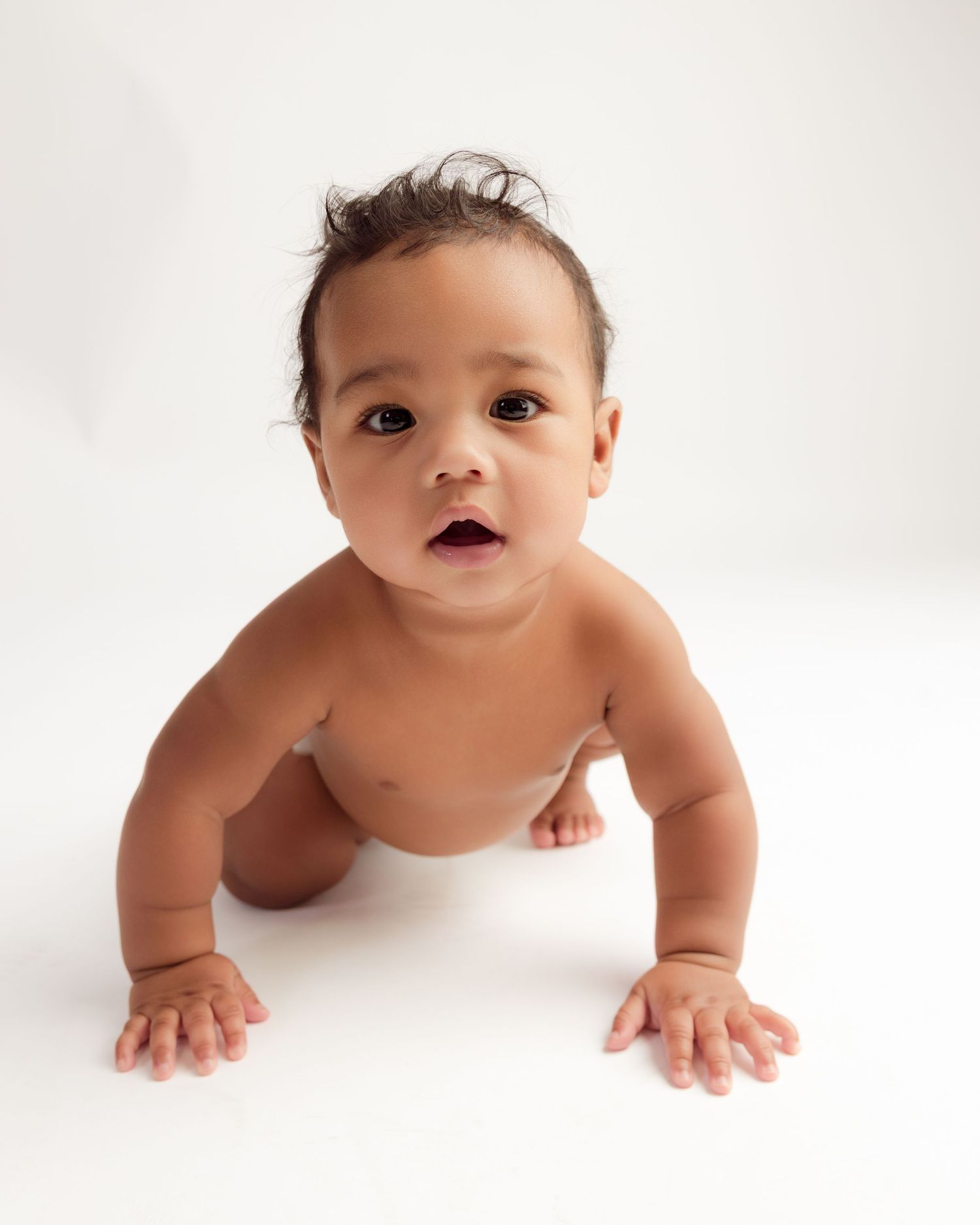 Adorable baby in a diaper crawling on a white background, looking up curiously with dark hair and bright eyes.