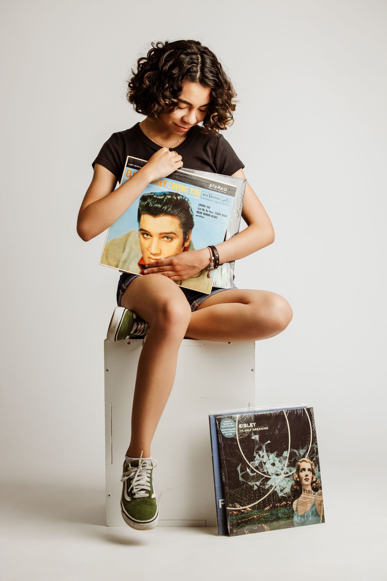 A person in casual attire sits on a white box while holding and examining vinyl record albums in a minimalist setting.
