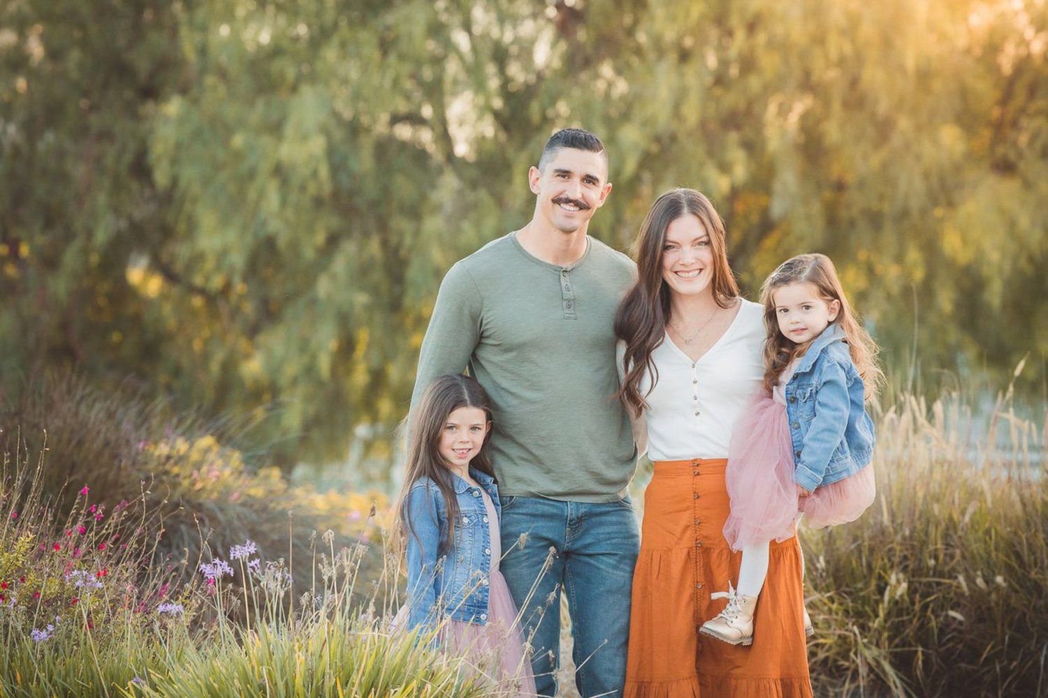 Happy family of four posing outdoors in golden hour light, parents with two young daughters in a lush green park setting.