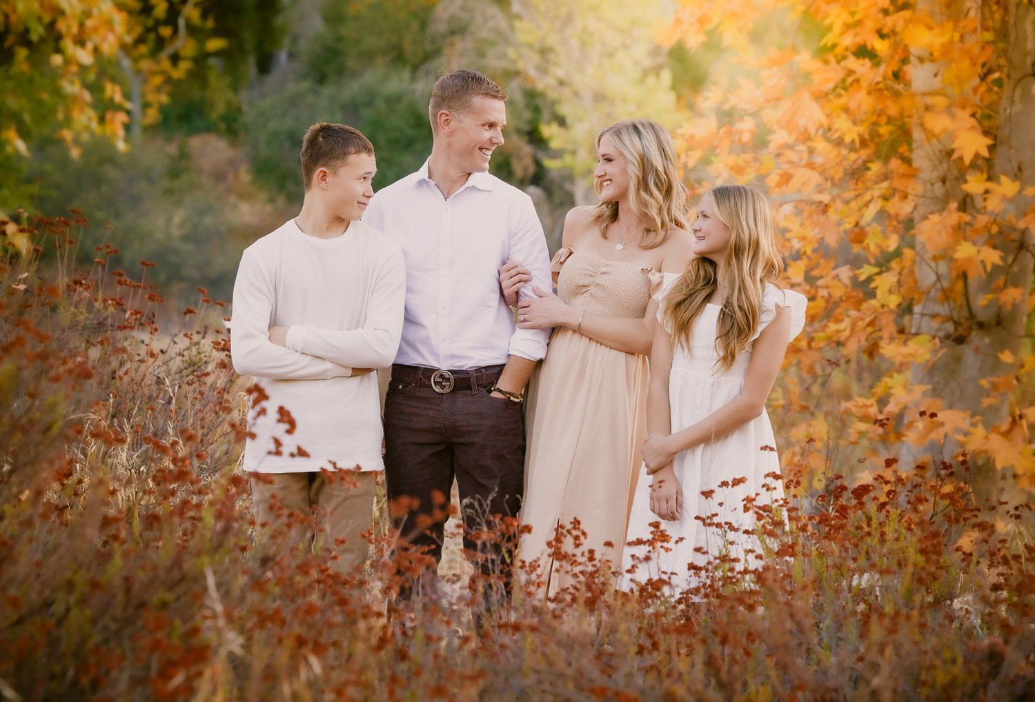 Family of four smiling together in a golden autumn forest, wearing white and neutral tones for a fall portrait.