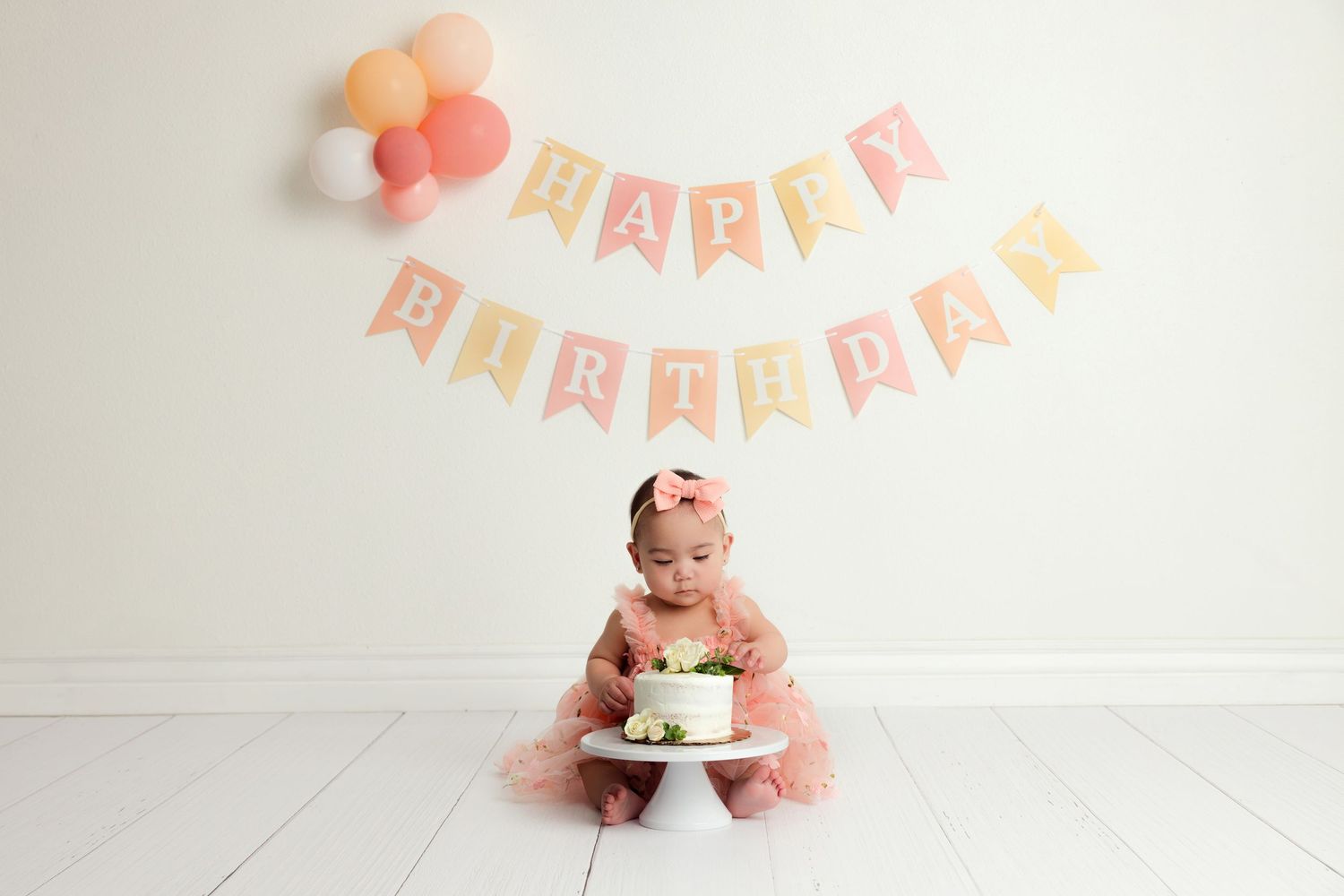 Baby girl in pink tutu smashes first birthday cake under Happy Birthday banner with pastel balloons.