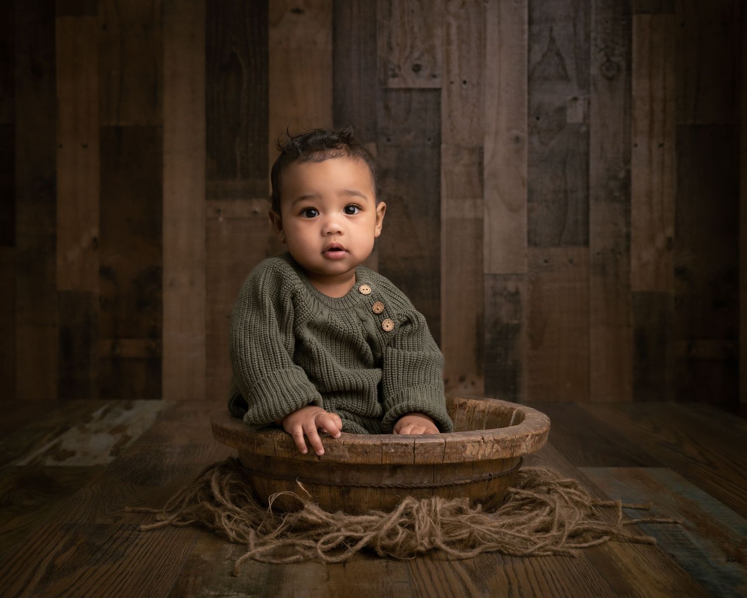 Adorable baby in green knit romper sitting in wooden bowl on rustic wood floor backdrop.