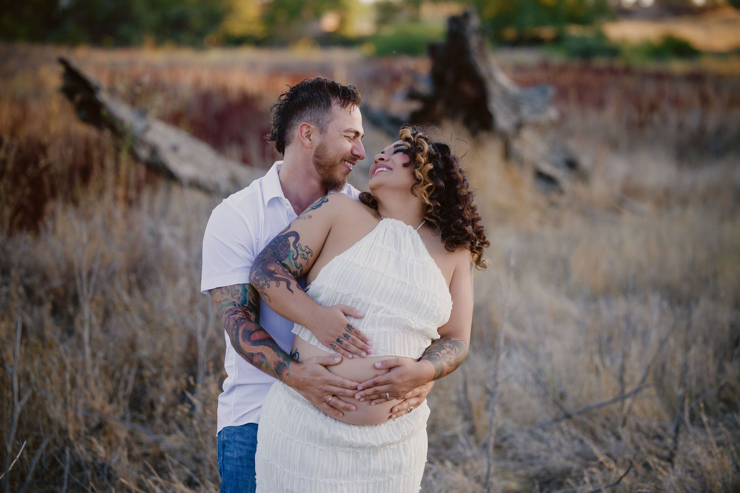 Couple embracing in golden field, pregnant woman in white dress, tattooed man kissing her cheek during maternity photo shoot.