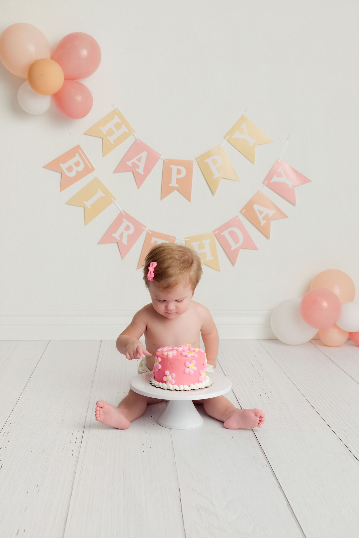 Baby girl sitting on white floor smashing pink birthday cake under Happy Birthday banner with pink and peach balloons.
