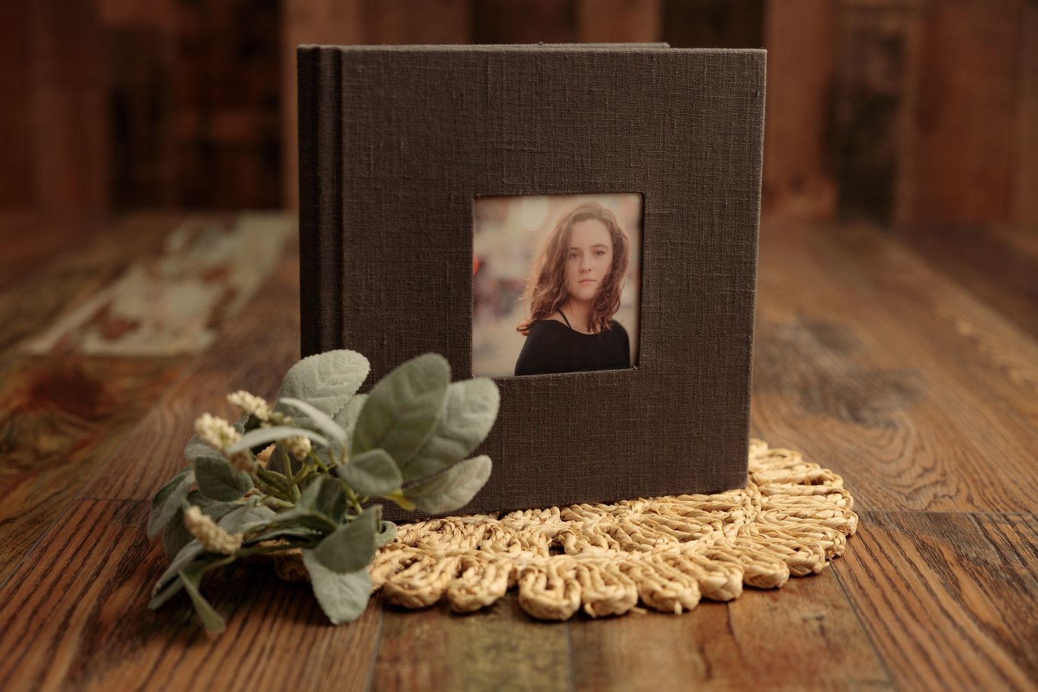 A vintage-style photo album rests on a rustic wooden table with eucalyptus leaves and scattered photographs.