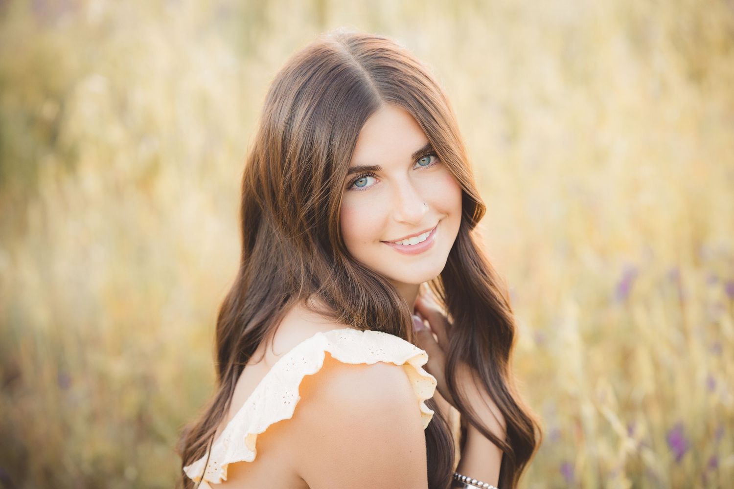 Young woman with long brown hair smiling over her shoulder in a sunlit golden field, wearing a ruffled top.