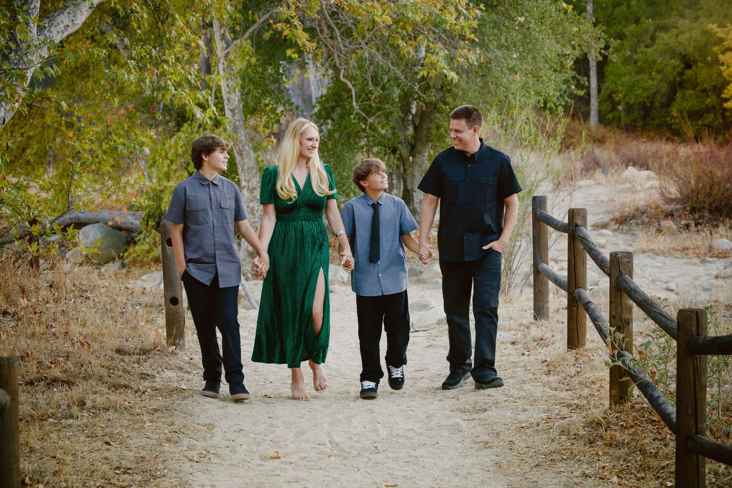 Family of four walking hand in hand along a dirt path beside a wooden fence, surrounded by trees in warm, golden outdoor light.