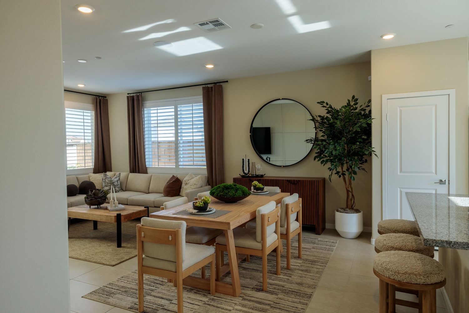 Modern open concept living room with beige sectional sofa, wooden dining table, and large round mirror on warm toned walls.