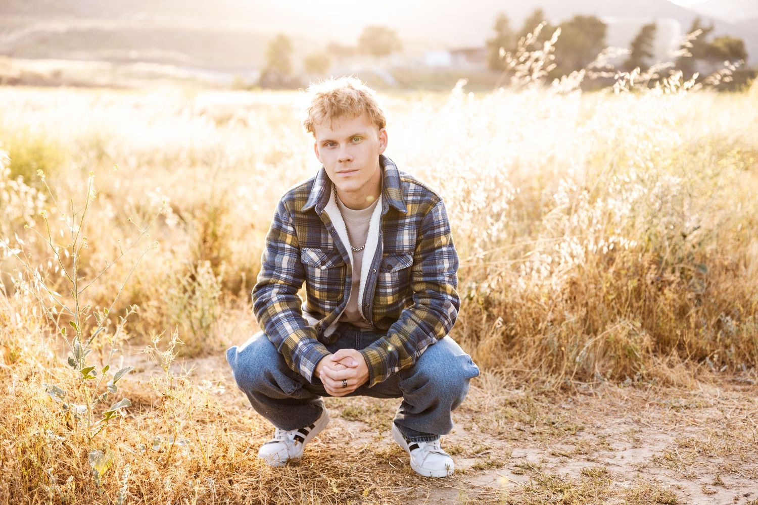 Young man in plaid shirt crouching in golden field during sunset for senior portrait photo.