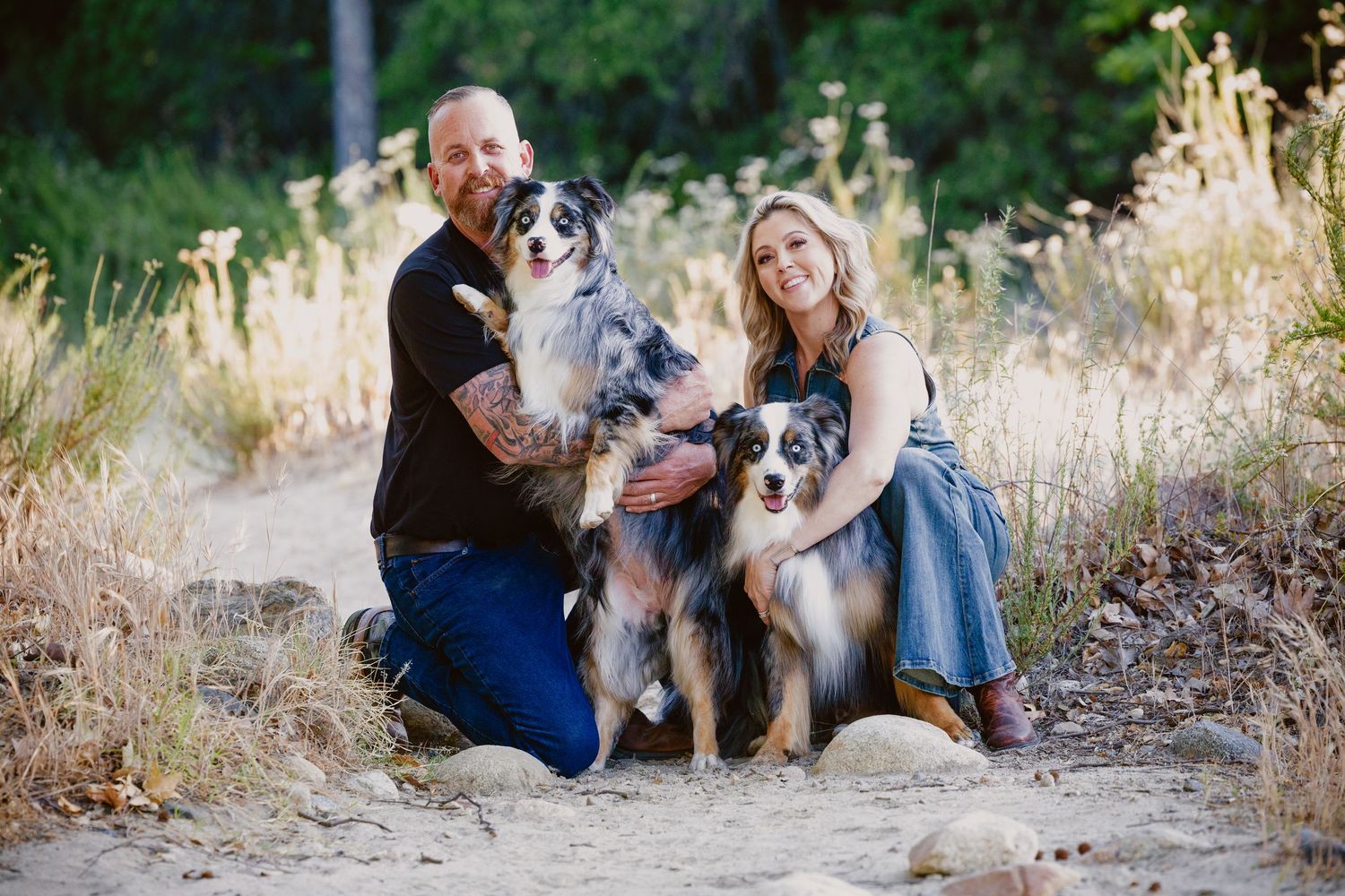 A smiling couple kneels on a sandy trail with two Australian Shepherd dogs in a sunny outdoor setting.