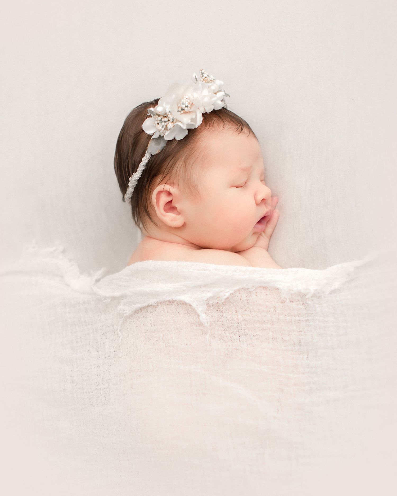 A peaceful sleeping newborn baby swaddled in white fabric with a delicate floral headband accessory.