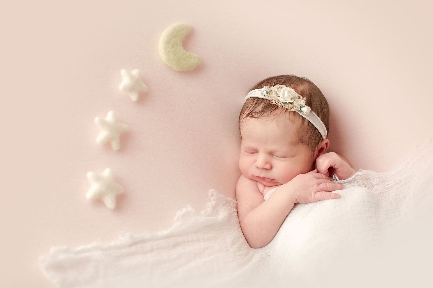 A sleeping newborn baby wrapped in pink fabric with delicate floral decorations around them in a soft studio setting.