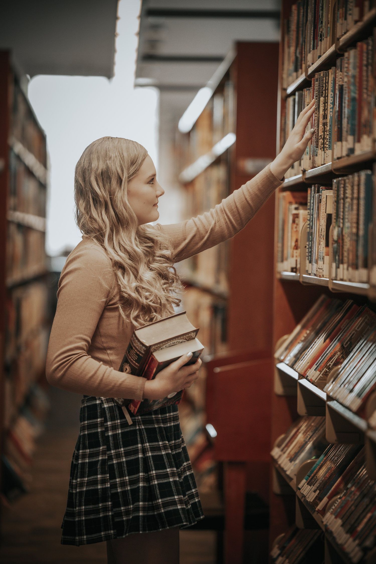 Blonde woman in plaid skirt browsing library bookshelves while holding a stack of books.