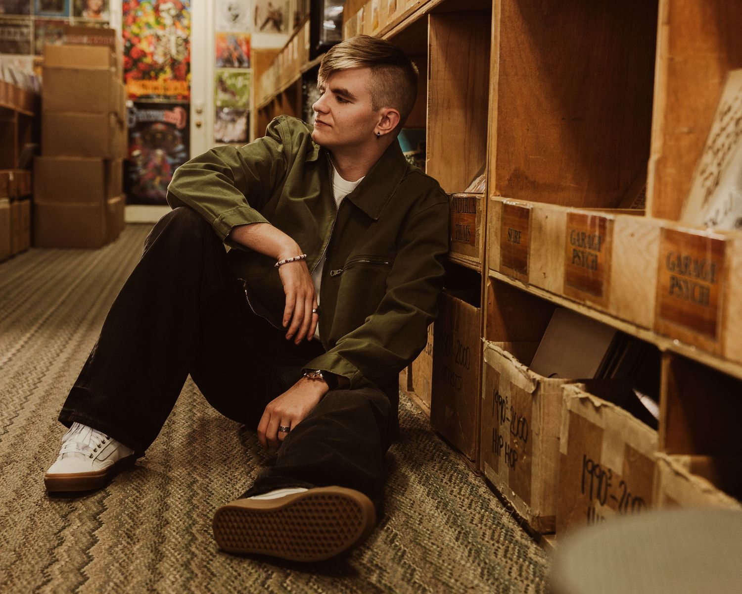 A person in casual attire sits on the floor against wooden shelving units in what appears to be a library or bookstore.