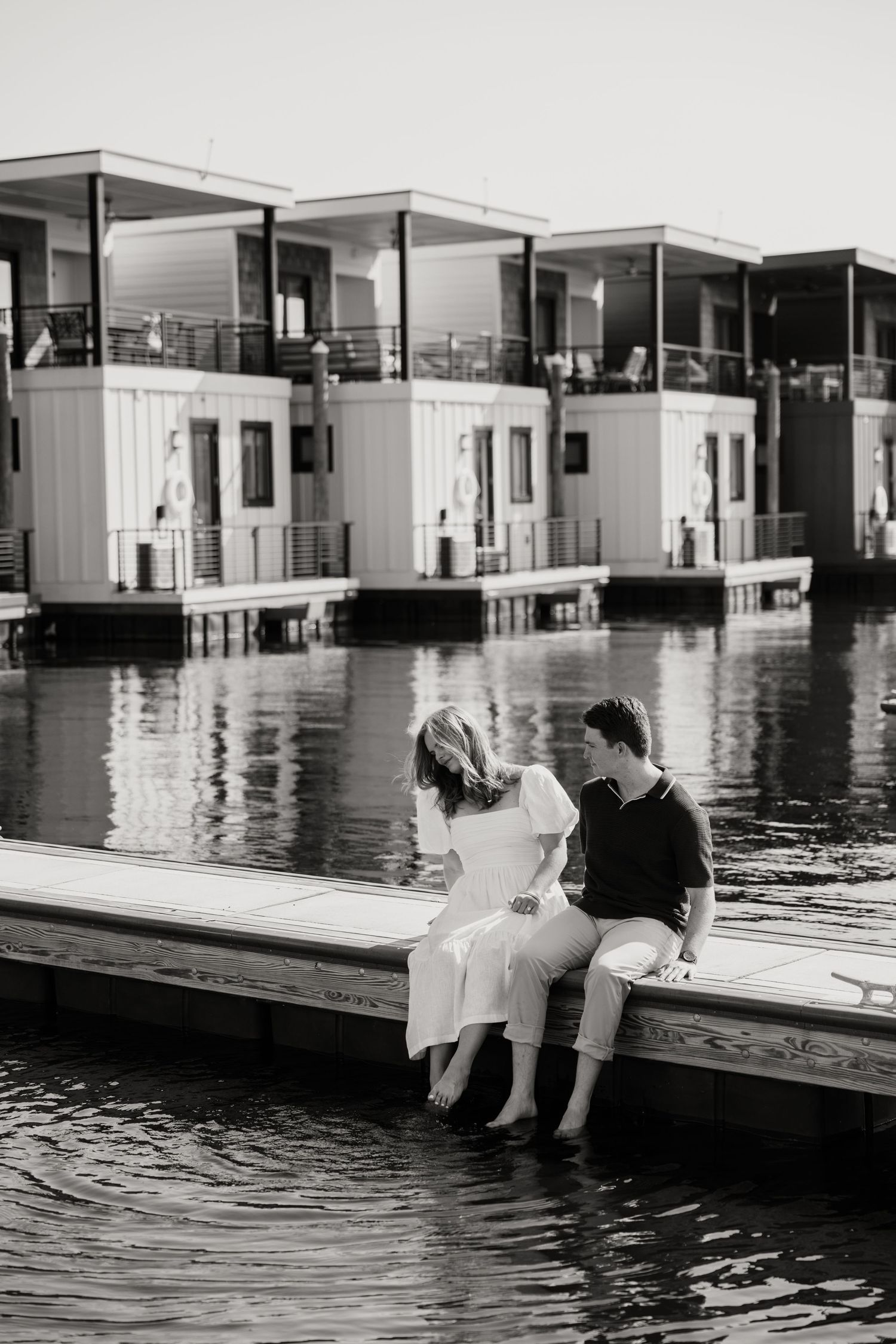 A couple sits at the edge of a modern pool deck with their feet dangling in the water at sunset.