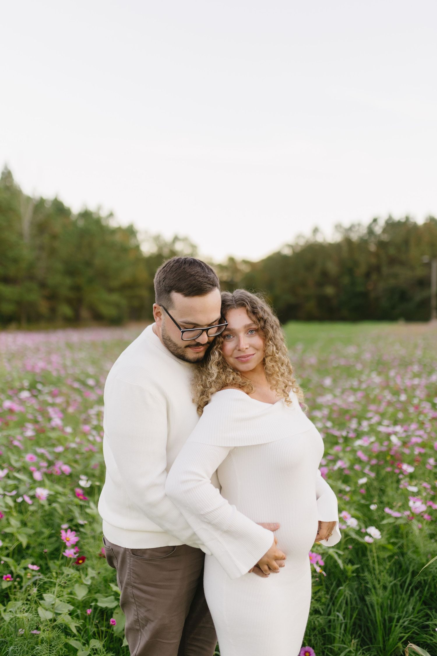 A couple embraces in a field of pink wildflowers, wearing matching white sweaters against a natural backdrop.