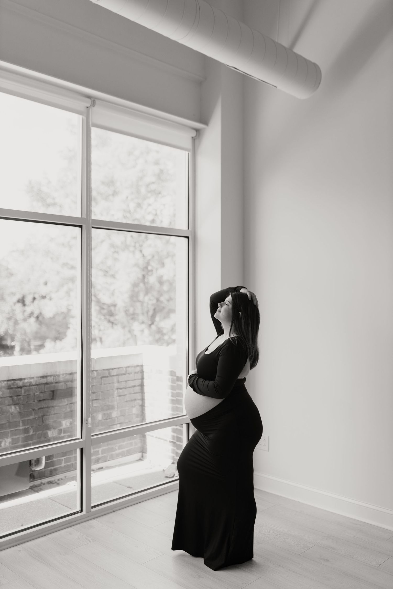 Silhouette of pregnant woman in black dress gazing out large window.