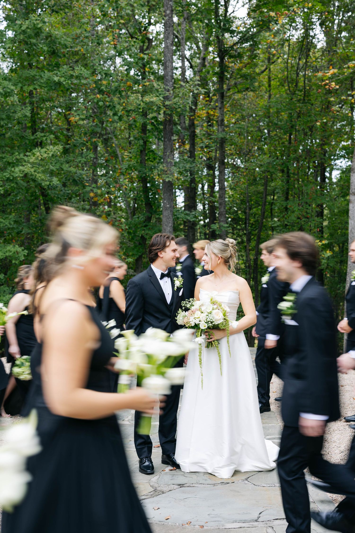 A wedding party in black and white attire gathers on a stone path surrounded by lush green forest trees.