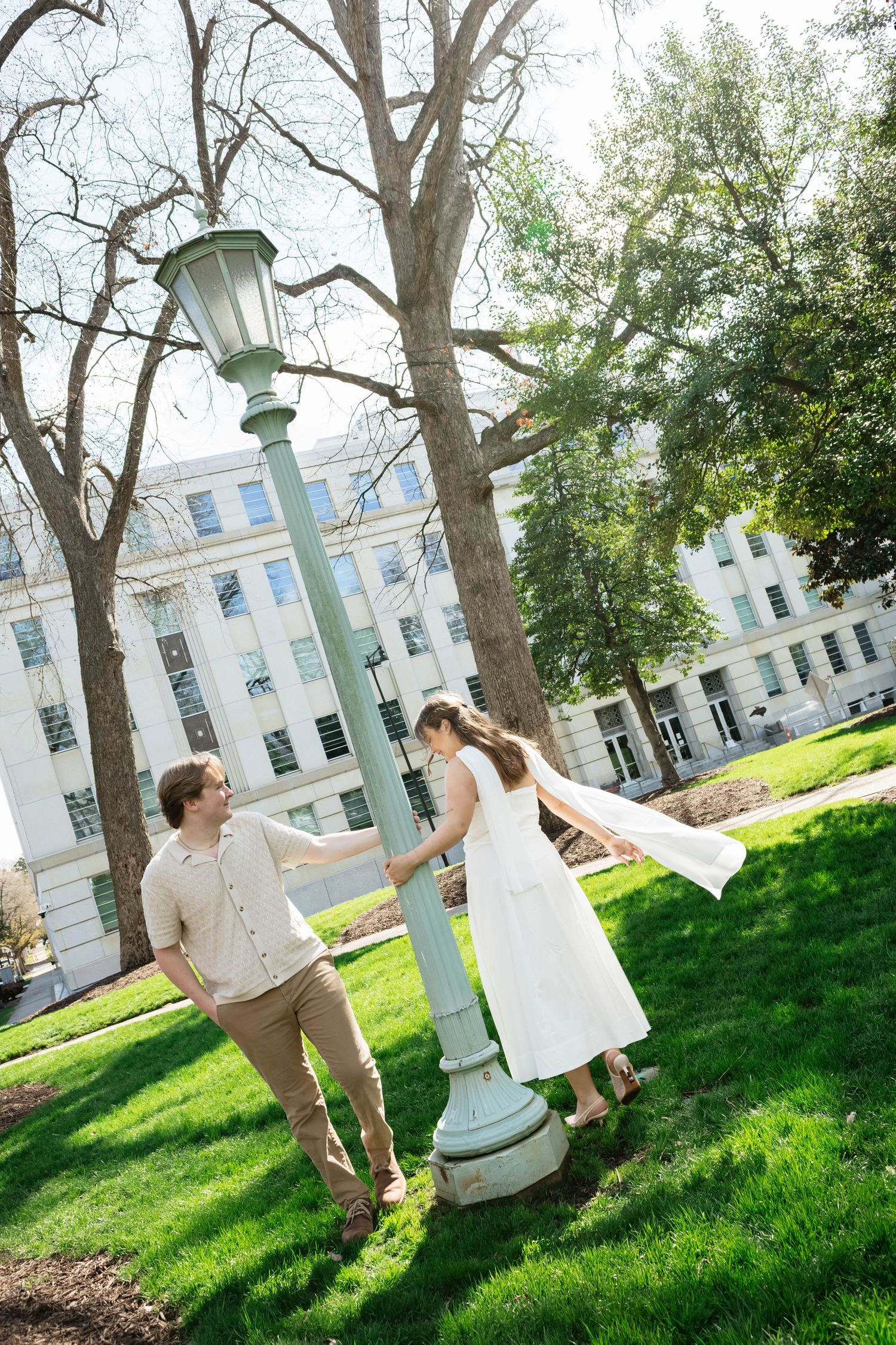 A couple playfully interacts near a vintage lamppost on a sunny day with historic buildings in the background.