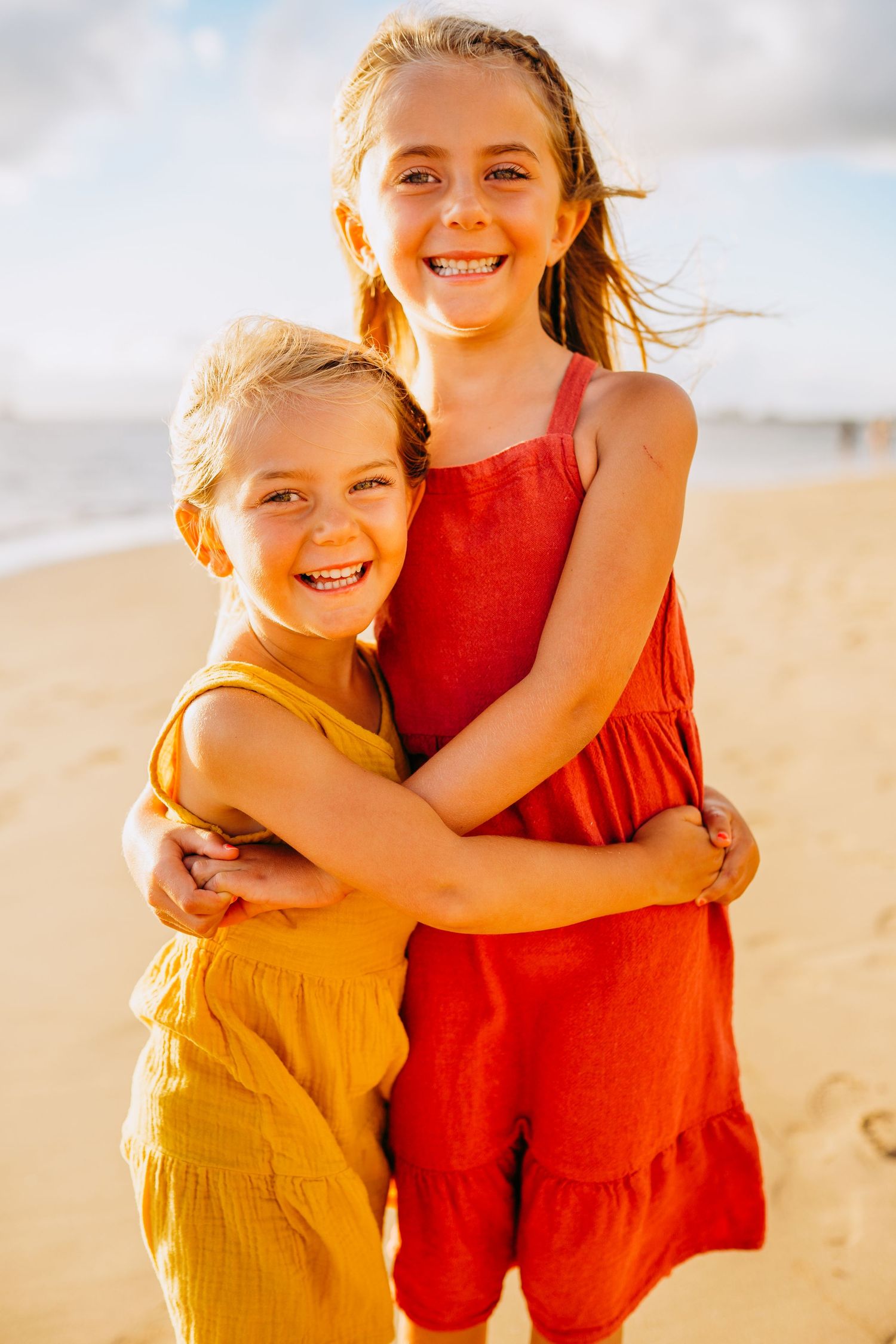 Two young children in yellow and red outfits embrace and laugh joyfully on a sunny beach.