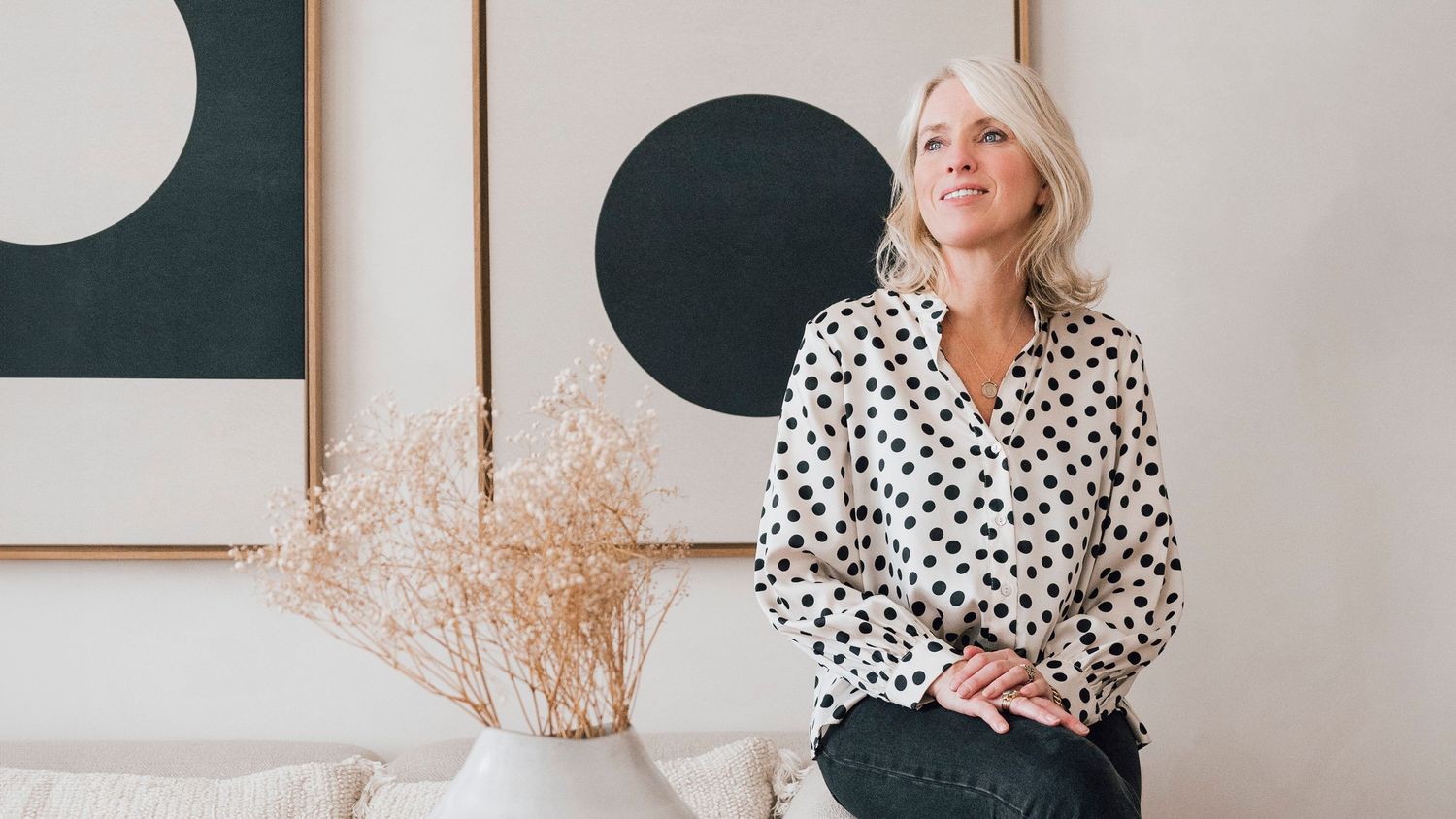 Vrouwelijke coach zit ontspannen in een lichte praktijkruimte met kunst aan de wand, kijkend naar buiten. Personal branding stories space.