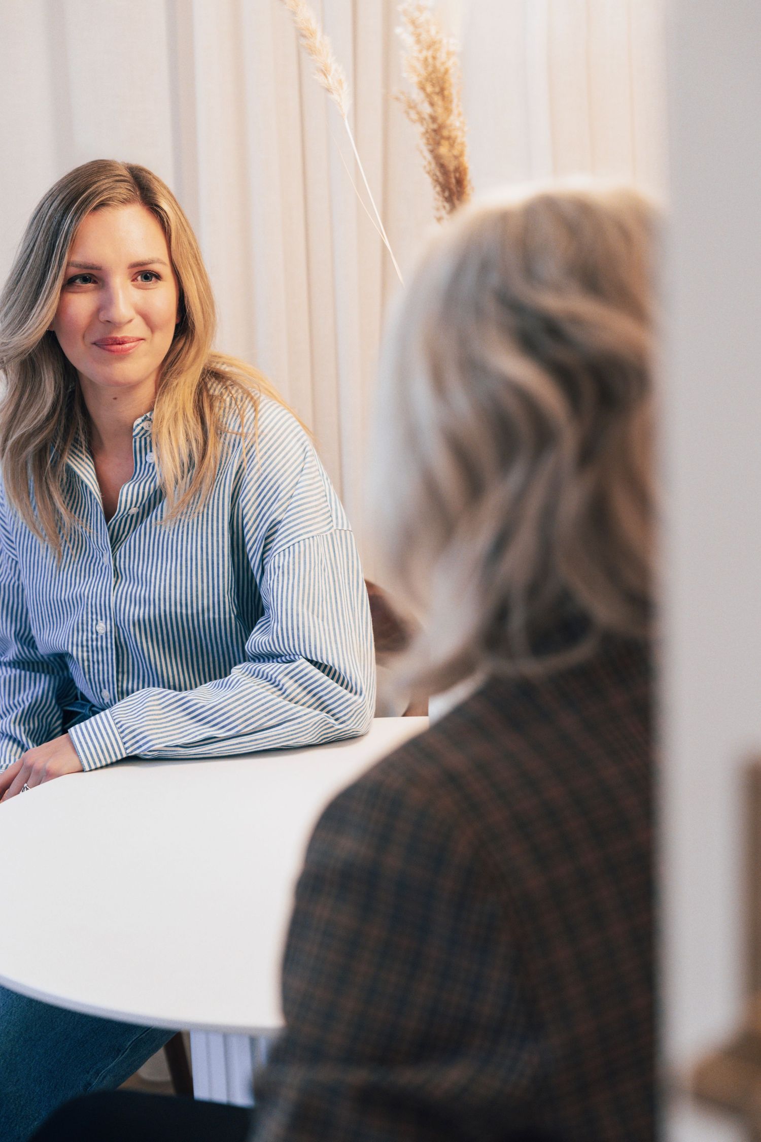 Vrouwelijke coach zit ontspannen in een lichte praktijkruimte, in gesprek met client