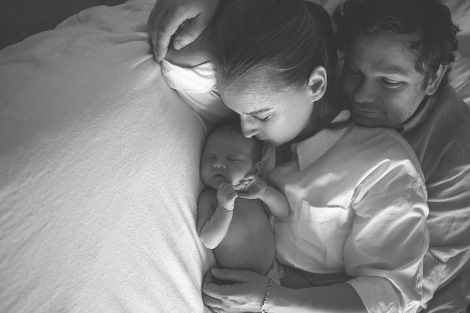 Family photography session in Prestwich, capturing a family of three lying on the bed together looking lovingly at their newborn baby.