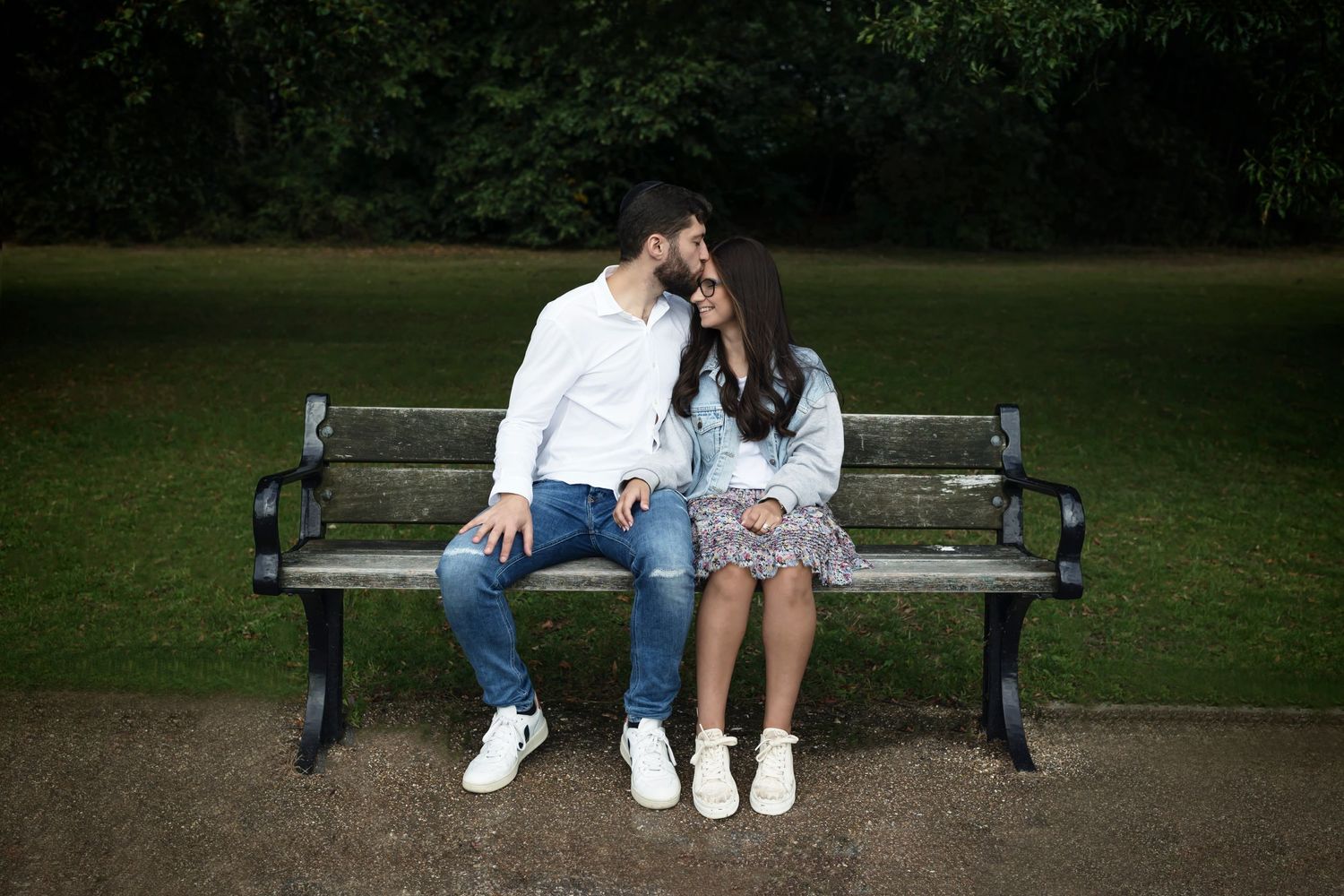 Couple sitting on a park bench, with the husband gently kissing his wife on the forehead.  Loving moment during a photo session in Prestwich