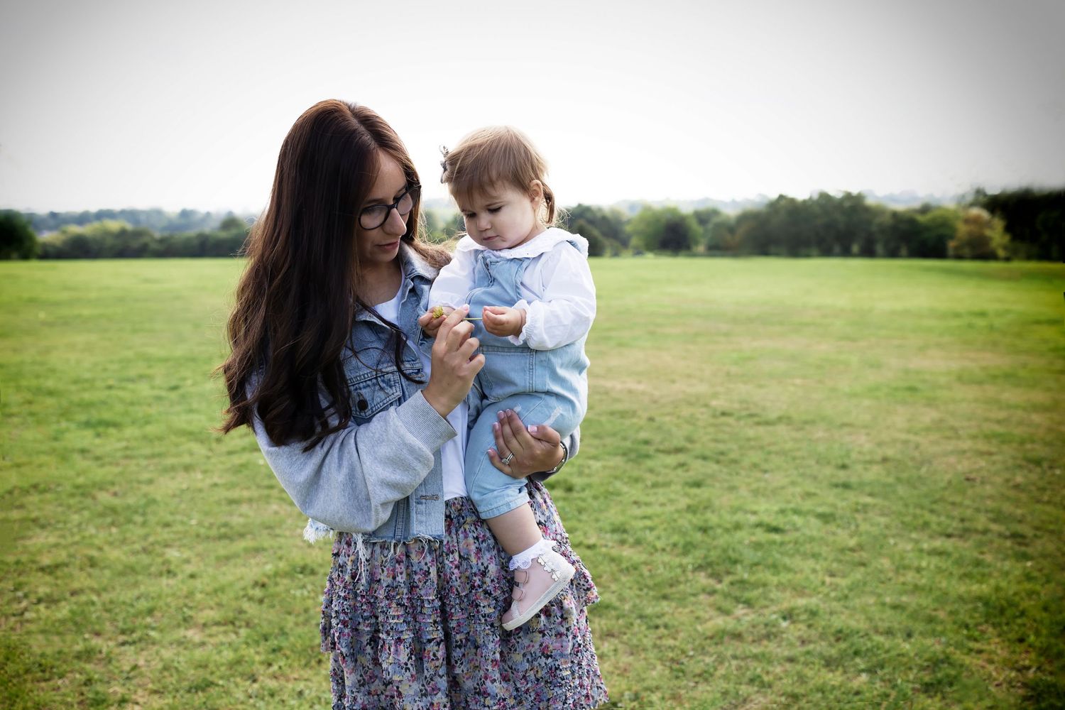 Lifestyle family photograph of a mum holding her  daughter in a local park in Prestwich, Manchester. Mum and daughter looking at a flower.
