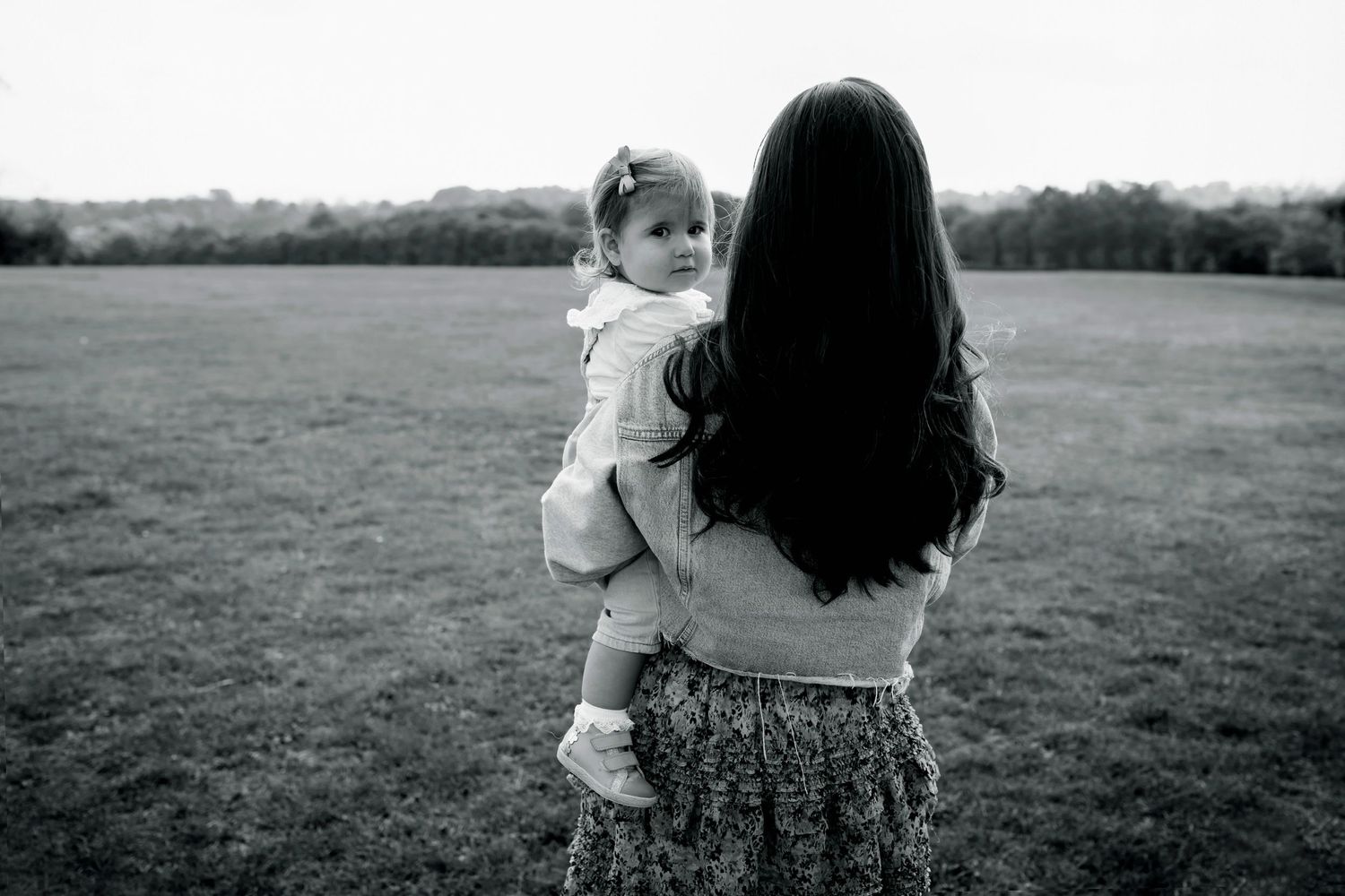 A mother with her back to the camera, holding her child who is peering over her shoulder, in a beautiful park setting in Manchester