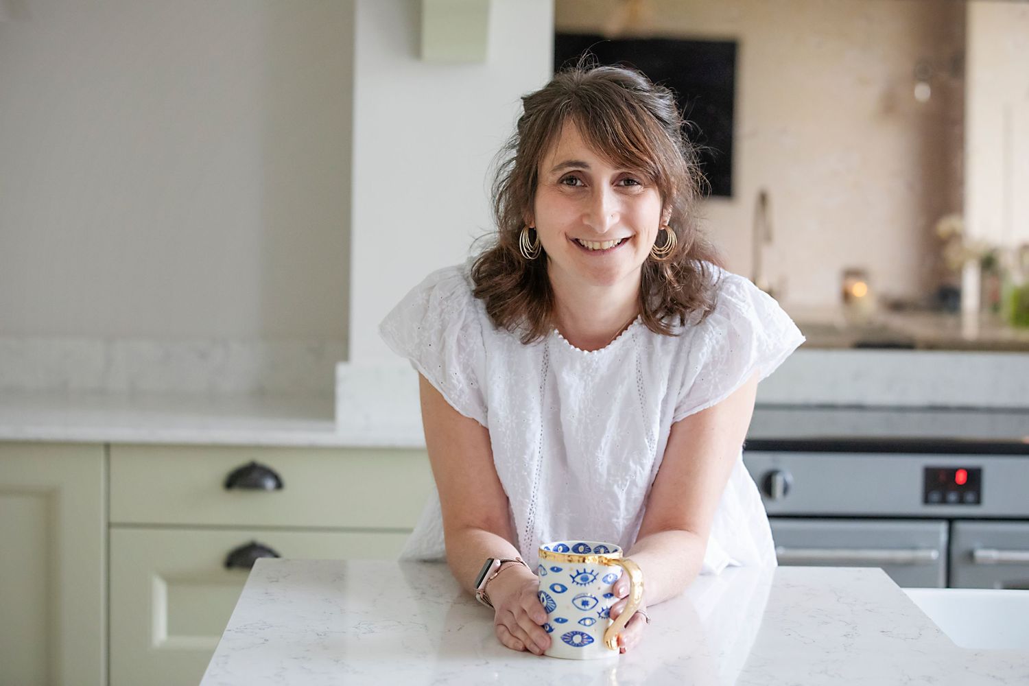 A woman holiding a cup of tea in her kitchen in Prestwich, looking at the camera with a warm smile during a personal branding photo session.