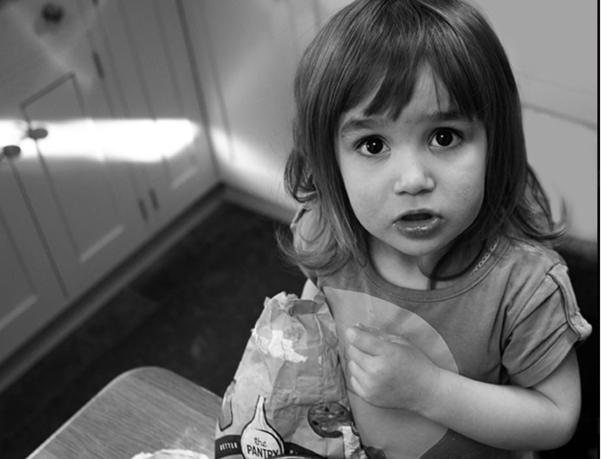 Young girl with flour-covered hands, looking up at the camera while baking in the kitchen during a family photo session in Whitefield.