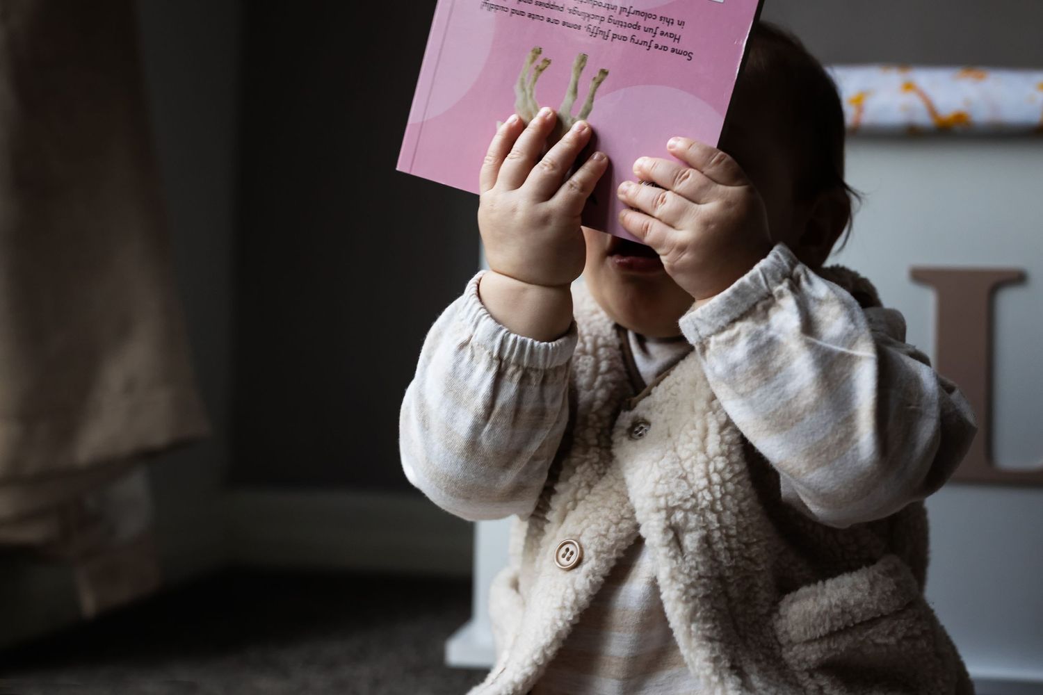 A young baby holds up a book in both hands covering his face. Prestwich, Manchester, UK