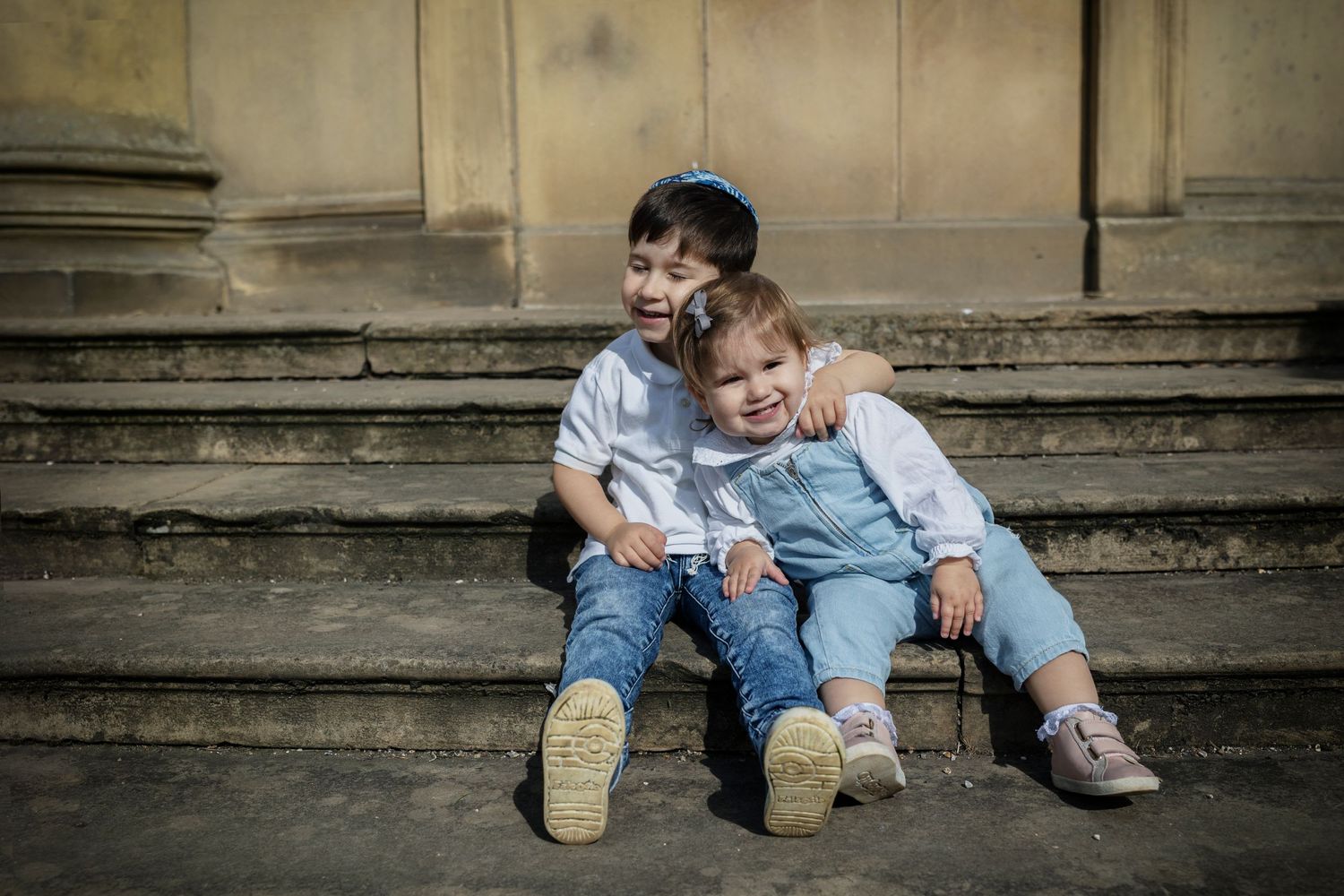 Two siblings sitting together on stone steps in a park and cuddling during a family photo session in Prestwich