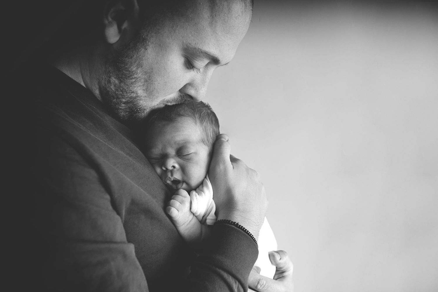 Black and white photography of dad lovingly kissing his newborn baby in a soft, serene room in Prestwich