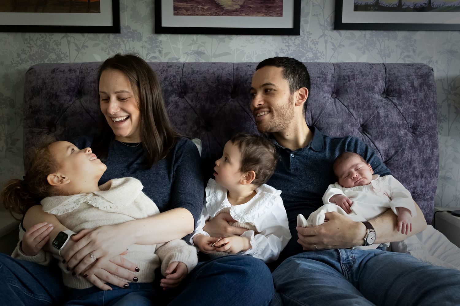 A family of five on the bed, with a newborn sibling in their parents' arms, smiling together during a family photo session in Whitefield