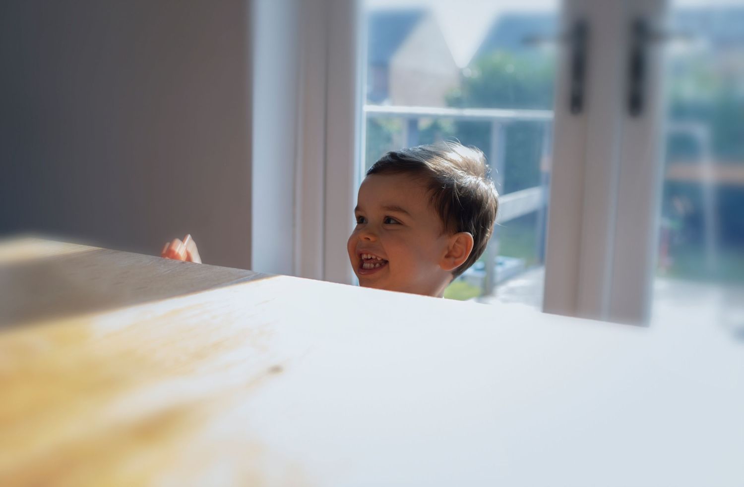 A playful boy peeking up from behind the kitchen table, smiling mischievously during a family photo session in Prestwich, Manchester