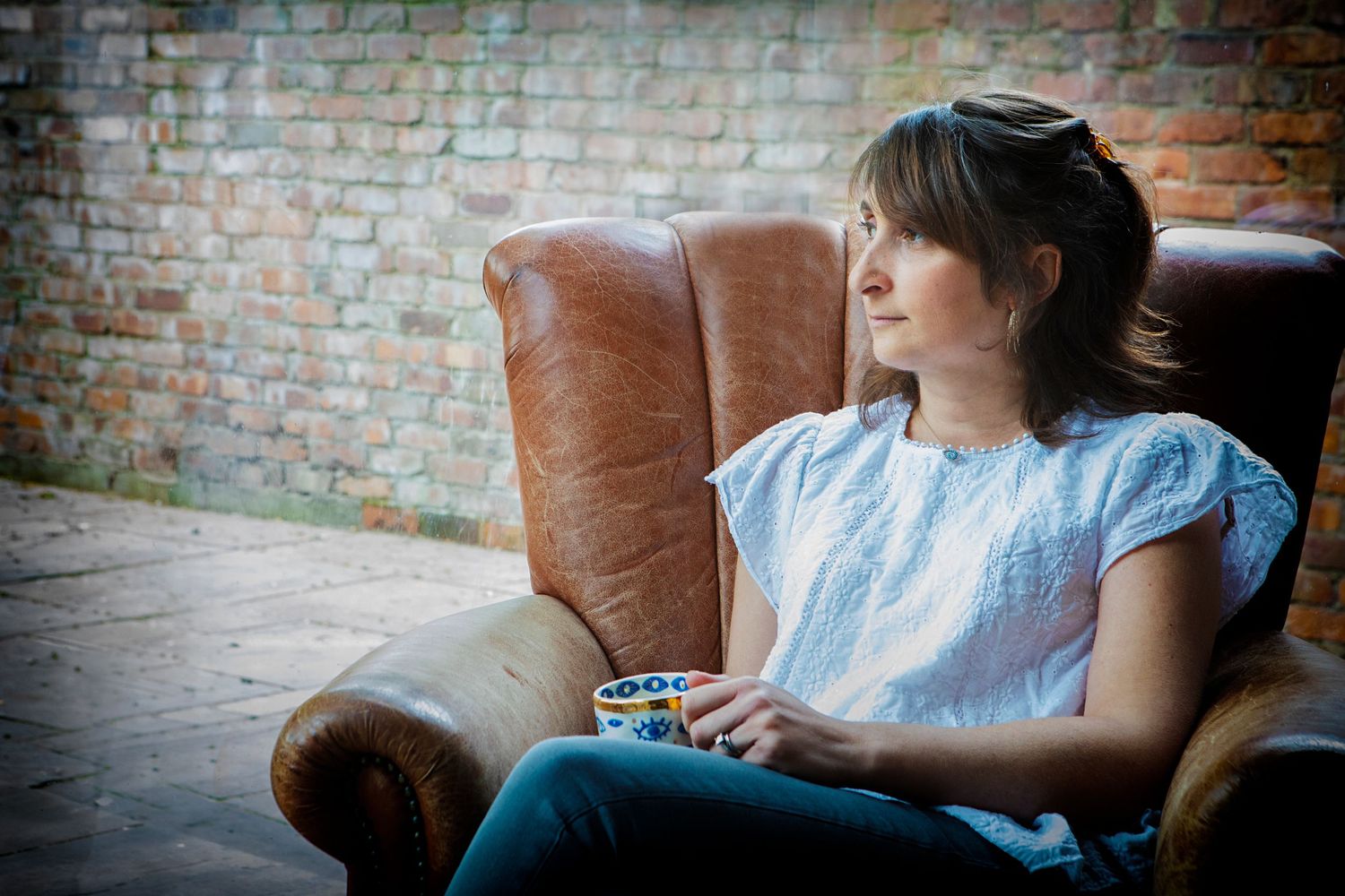 Woman sitting on a comfy chair in her home in Prestwich, looking out the window and deep in thought during a personal branding photo session