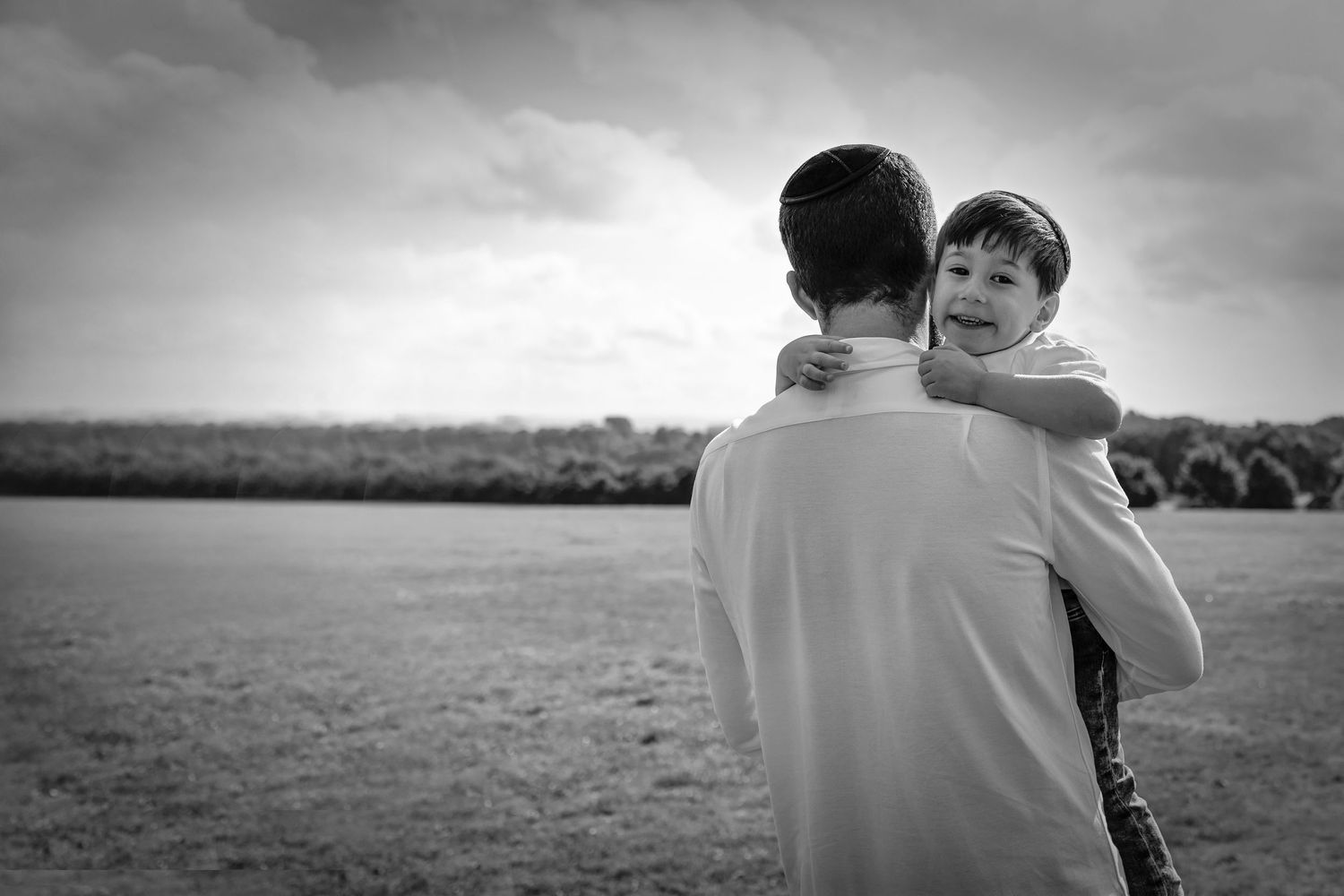 Black and white photo of a dad holding his son over his shoulder in a park. Dad's back is to  camera and the son looks at camera. Manchester