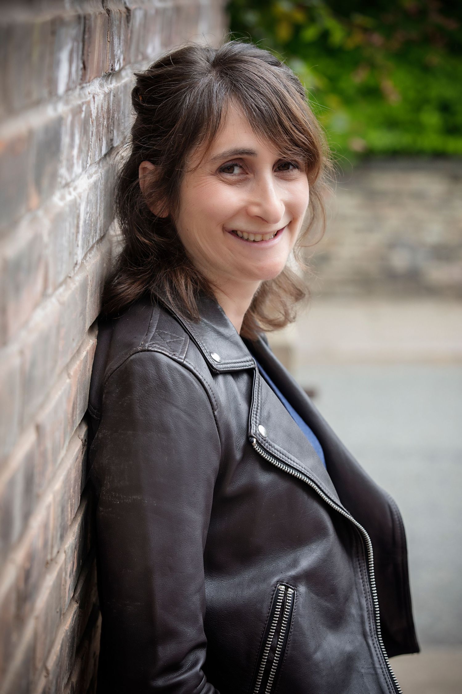 woman smiling and leaning with her back against a brick wall in her home in Prestwich, captured during a personal branding photo session.