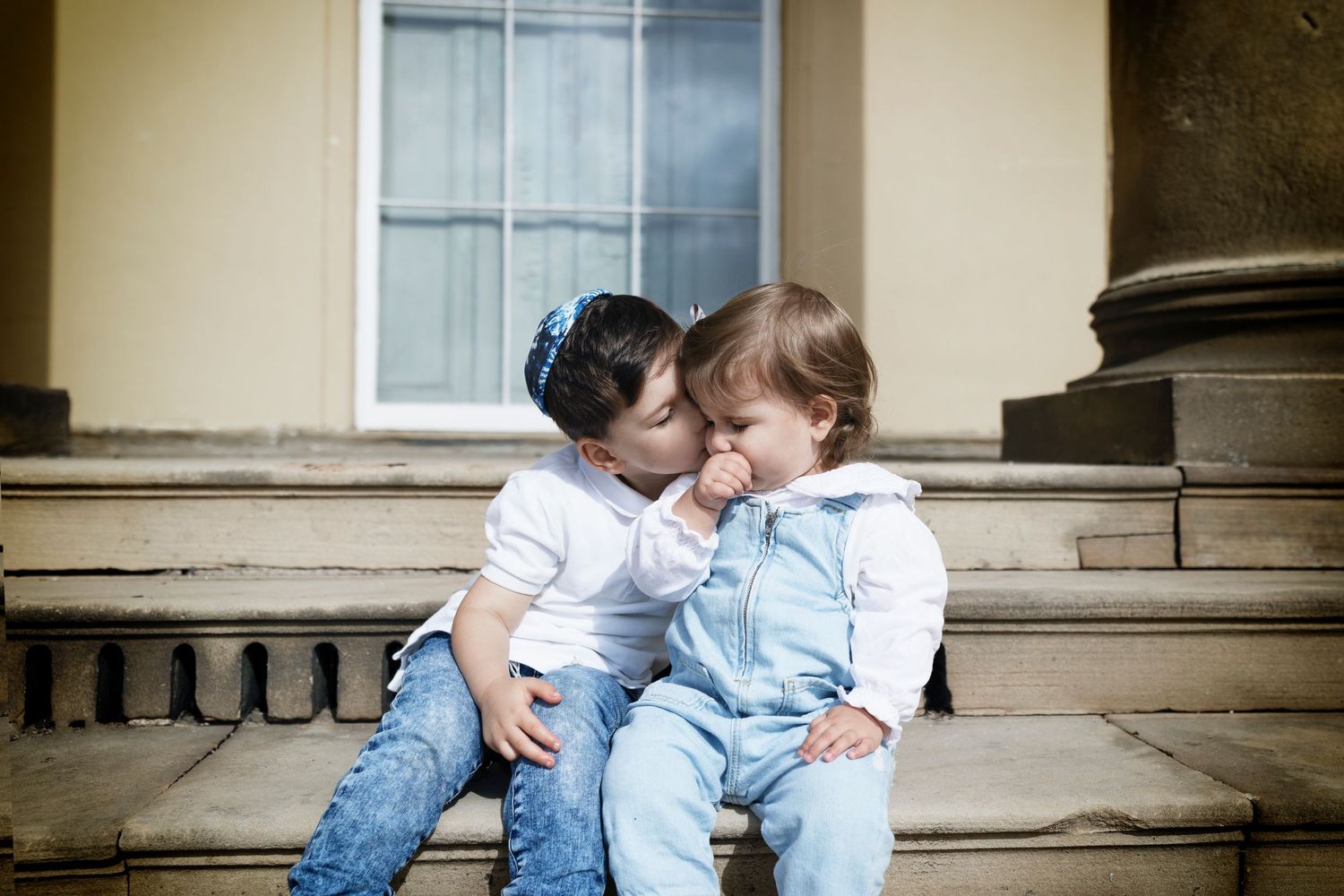 Young brother gives his sister a kiss on the cheek during a family photo session in a local park in Prestwich