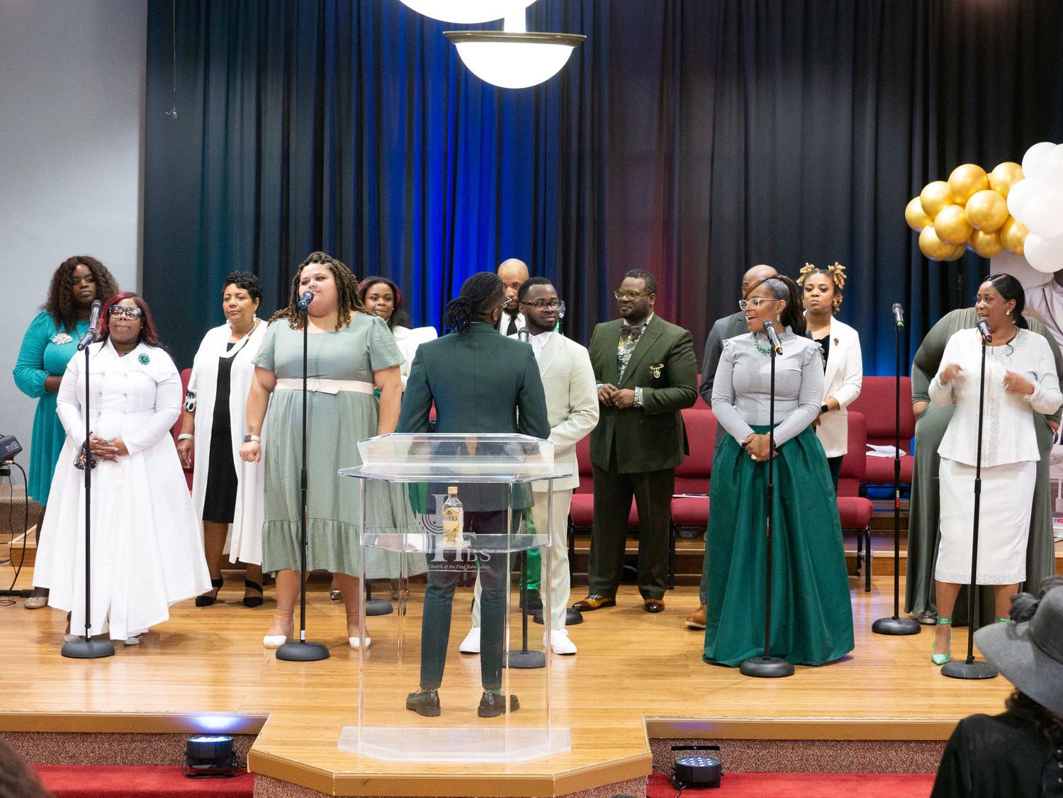 Gospel choir performs on stage with a clear podium and gold balloon decorations in the background.