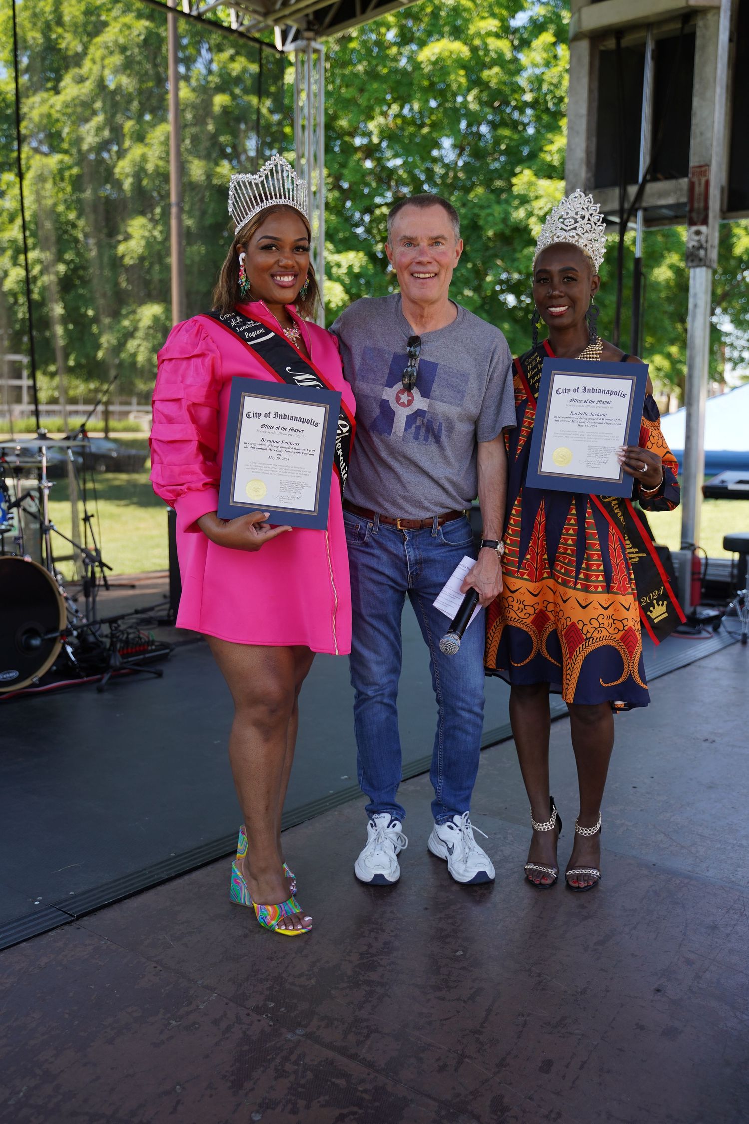 Two pageant winners in colorful outfits display their award certificates alongside a man in casual wear on stage.